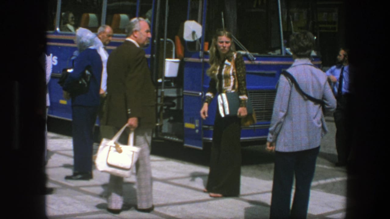 A woman in a patterned jacket stands near a blue bus while a man with a white handbag turns in the foreground.