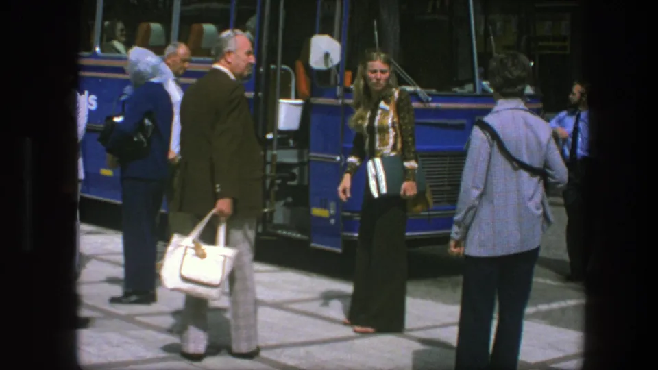 1975 Copenhagen street scene with a woman in patterned clothing near a blue bus and a man carrying a white handbag among waiting adults.