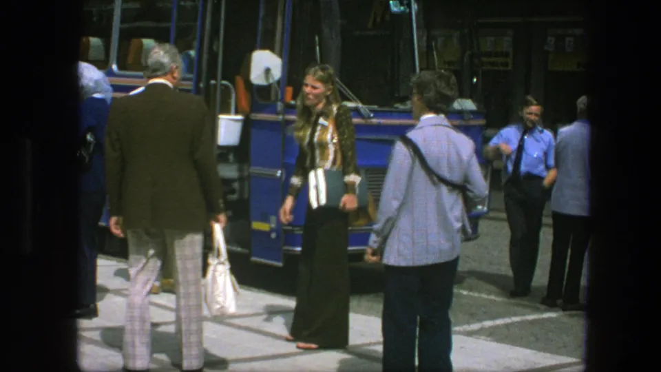 Copenhagen 1975 home-movie frame of adults near a blue bus, including a woman in flared pants and a man holding a white handbag.