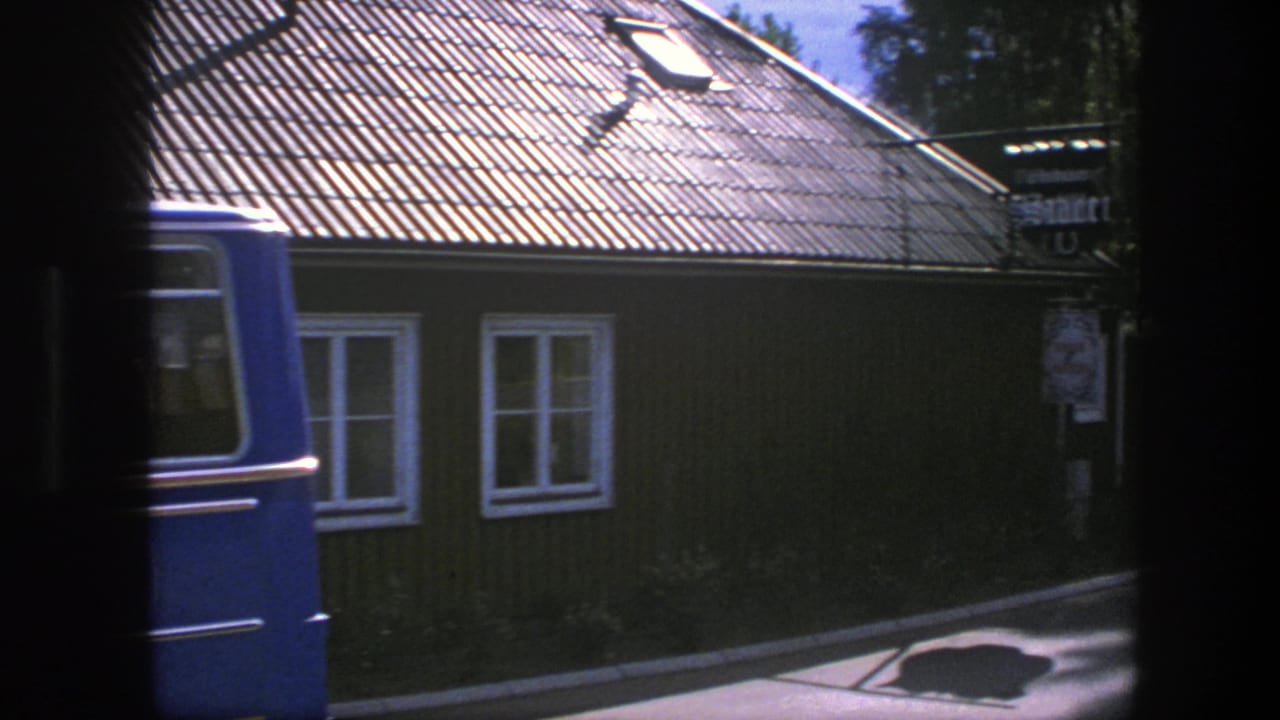 Blue van passing in front of the wooden building and white-framed windows.