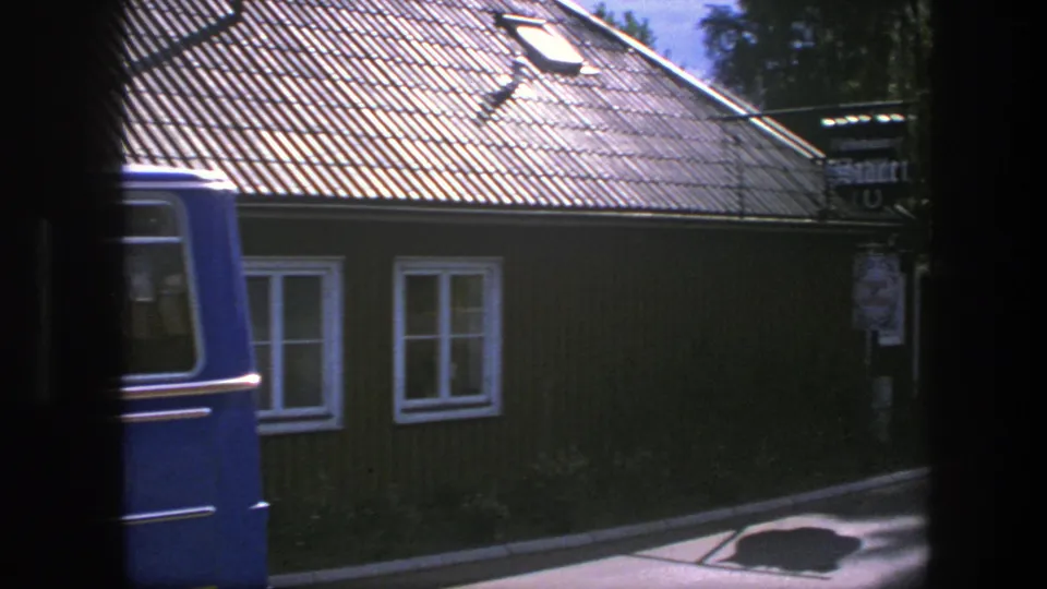 A blue van begins to pass across the 1975 Copenhagen view, partly covering the wooden building and roadside sign.