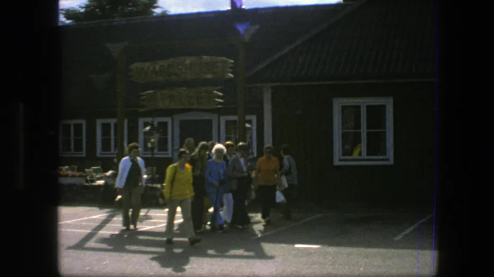 A group of tourists in 1975 Copenhagen walks toward a dark building with a wooden sign above the entrance area.