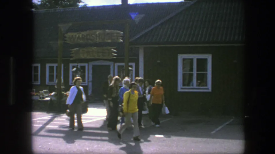 Tourists move across a sunlit paved area toward a dark building and wooden sign during a Copenhagen travel stop.