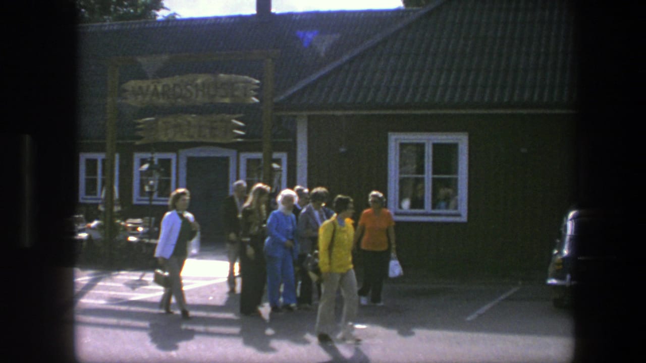 In Copenhagen, 1975, several people walk across pavement outside a dark green wooden building with a sign above the entrance.