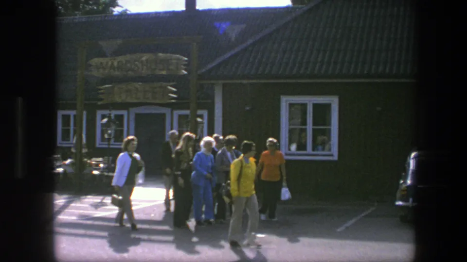 In Copenhagen, 1975, several people walk across pavement outside a dark green wooden building with a sign above the entrance.