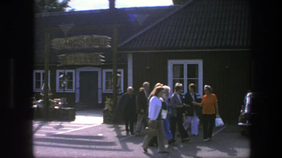 The 1975 Copenhagen home-movie frame shows adults in casual clothing moving past the building, with the sign and windows partly visible.