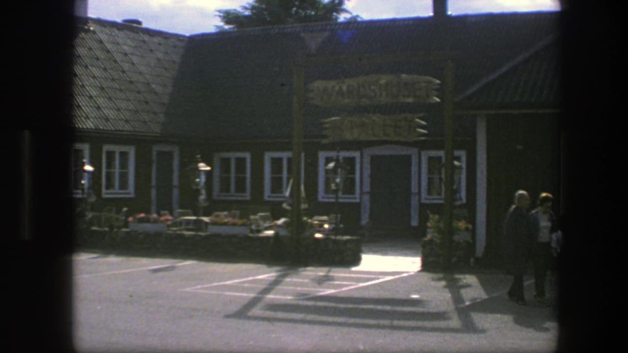 Brown building with white-trimmed windows, sign over the entrance, flower boxes, and pedestrians near the doorway.