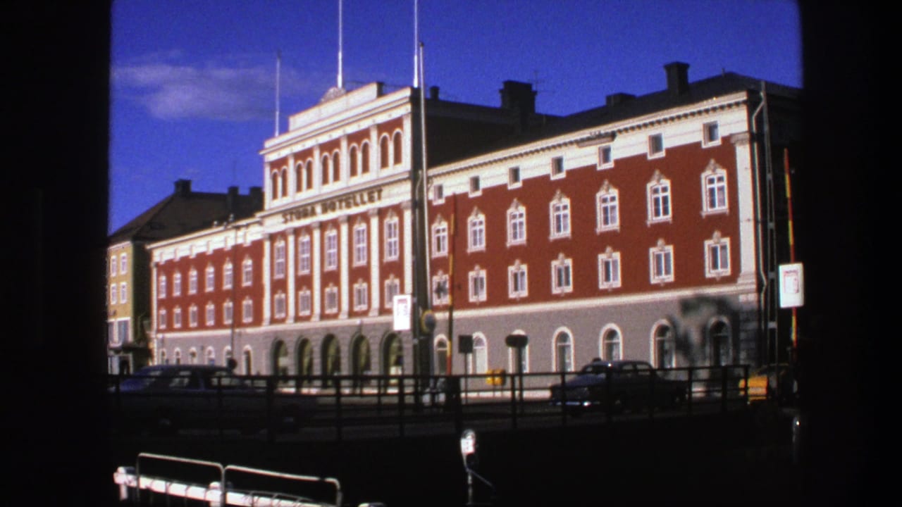Red and white building exterior with hotel sign, cars, railings, and sunny sky.