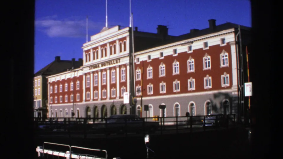 1975 Copenhagen home-movie frame showing the red brick and white-trimmed hotel exterior from across a street, with cars and possible pedestrians below.