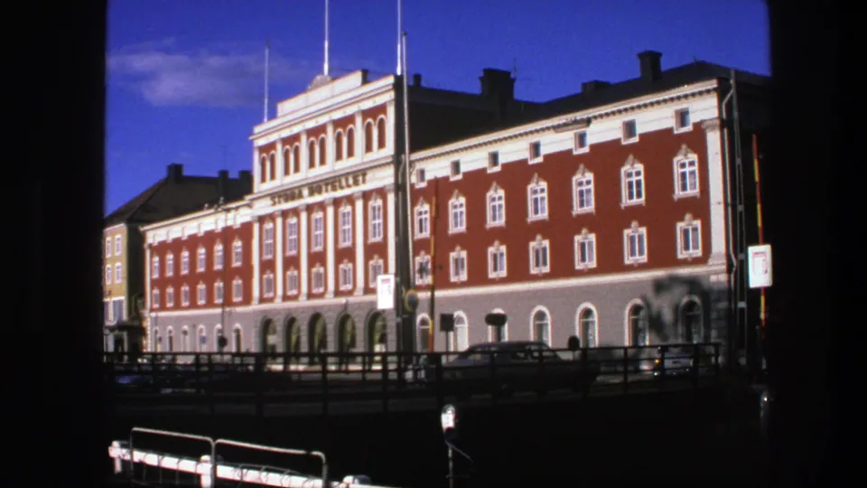 End frame of a 1975 Copenhagen exterior, holding on a red brick hotel-style facade with white trim, street railings, cars, and visible signage.