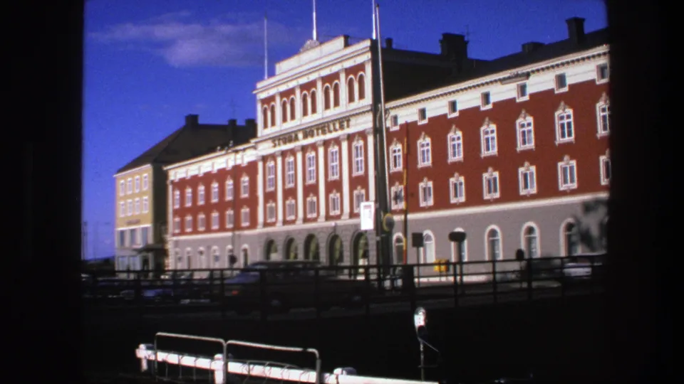1975 Copenhagen home-movie frame of a red and white hotel facade viewed across railings with traffic below.