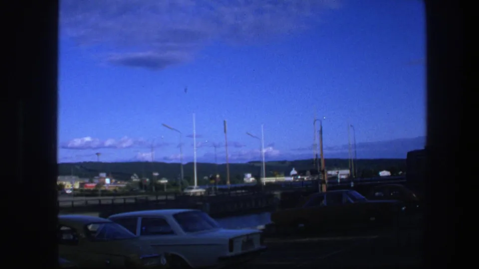 1975 Copenhagen 8mm view from near the hotel area, showing parked cars, boat masts, low buildings, distant hills, and a clear blue sky.