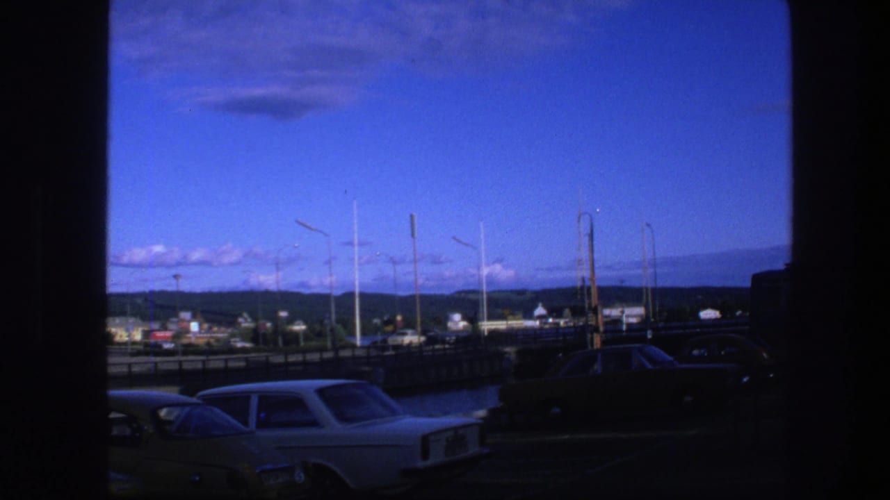 Classic cars near a roadway with tall lamps, low buildings, distant landscape, and blue sky.