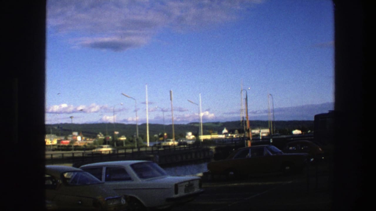 Parked classic cars beside a waterfront road with railings, distant buildings, hills, and blue sky.