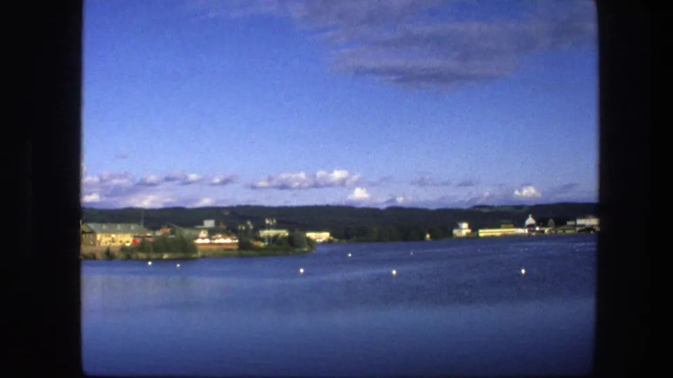 1975 Copenhagen waterfront frame showing open water with small buoys and a distant shoreline of trees, low buildings, and hills under a bright sky.