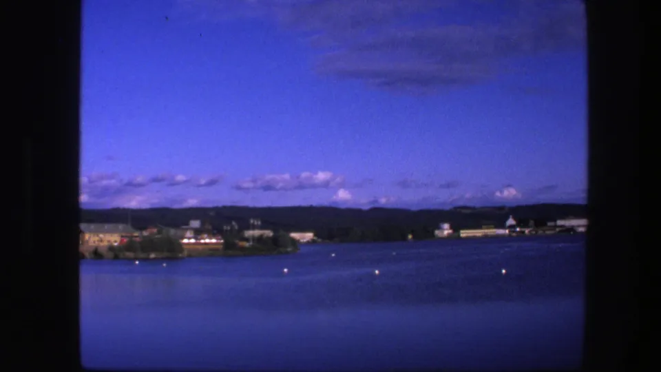 Blue-shifted 8mm frame of Copenhagen waterfront, with calm water, small buoys, distant shoreline buildings, trees, low hills, and clouds.