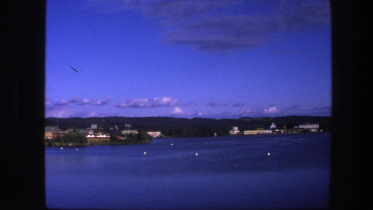 1975 Copenhagen lake view with calm blue water, small buoys, low shoreline buildings, distant hills, and 8mm frame edges.