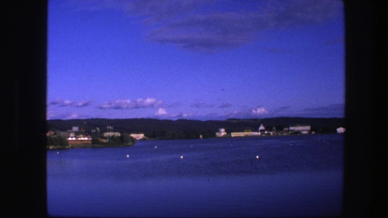 Calm lake with scattered buoys, distant buildings, wooded shore, hills, and blue sky.