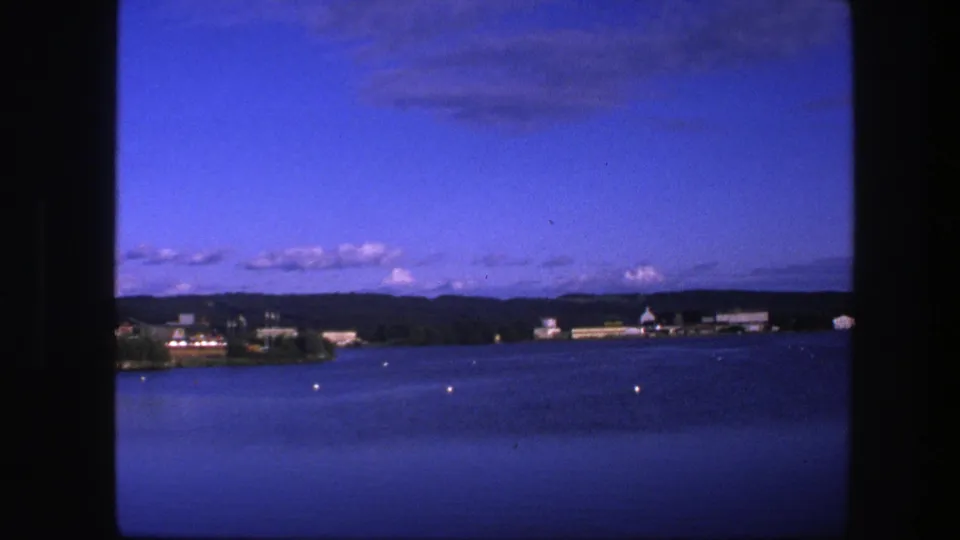 1975 Copenhagen waterfront scene with blue dusk sky, calm lake surface, small buoys, distant shore buildings, and hills.