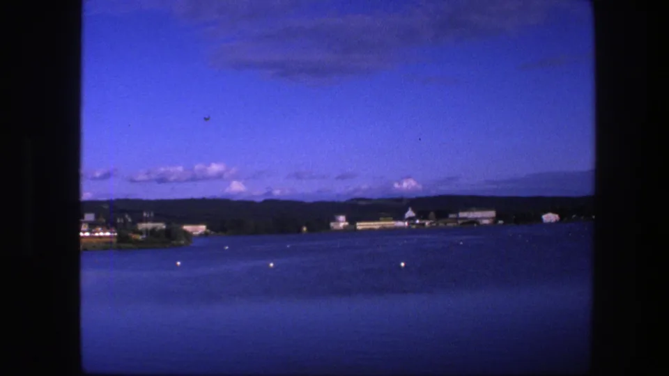 1975 Copenhagen lake view showing deep blue water, small buoys, distant cottages or dock buildings, clouds, and horizon hills.