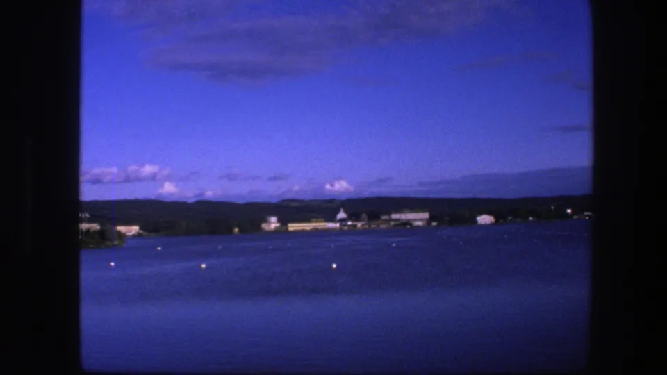 1975 Copenhagen lakeside view with calm water, buoy markers, low buildings along a tree-lined shore, distant hills, and 8mm borders.