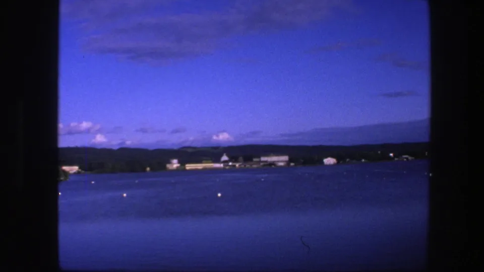 1975 8mm view from the sea toward Copenhagen, with calm water, scattered buoys, low shoreline buildings, and dark film borders.