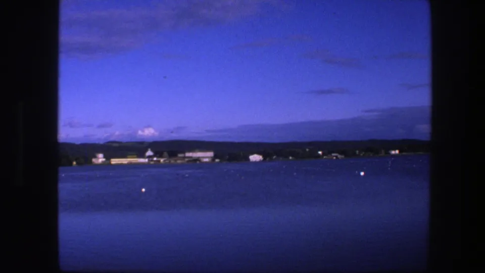 1975 home-movie frame looking over Copenhagen water toward a low shoreline, with buoys, buildings, and saturated blue 8mm color.