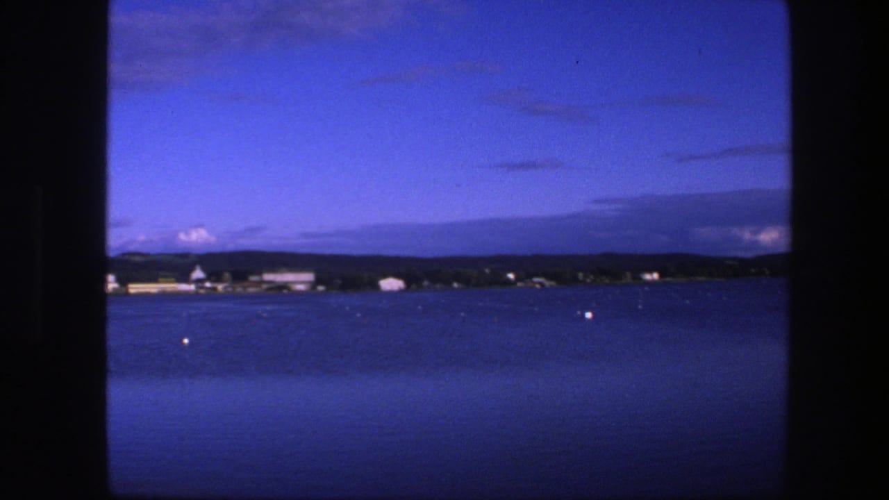 Blue water at dusk with a distant shoreline, low buildings, trees, clouds, and black 8mm frame borders.