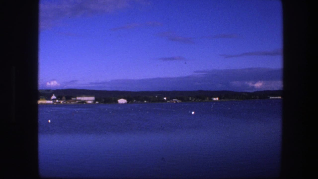 1975 Copenhagen-area home-movie frame with calm blue water, a low distant shore, pale waterfront structures, and dark clouds near the horizon.
