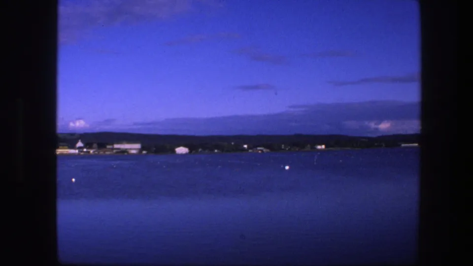 1975 Copenhagen-area home-movie frame with calm blue water, a low distant shore, pale waterfront structures, and dark clouds near the horizon.