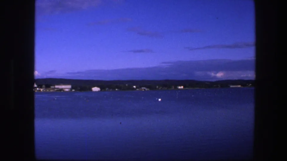 1975 Copenhagen-area 8mm view over dark blue water toward a low shore with small pale buildings, trees, and layered dusk clouds.