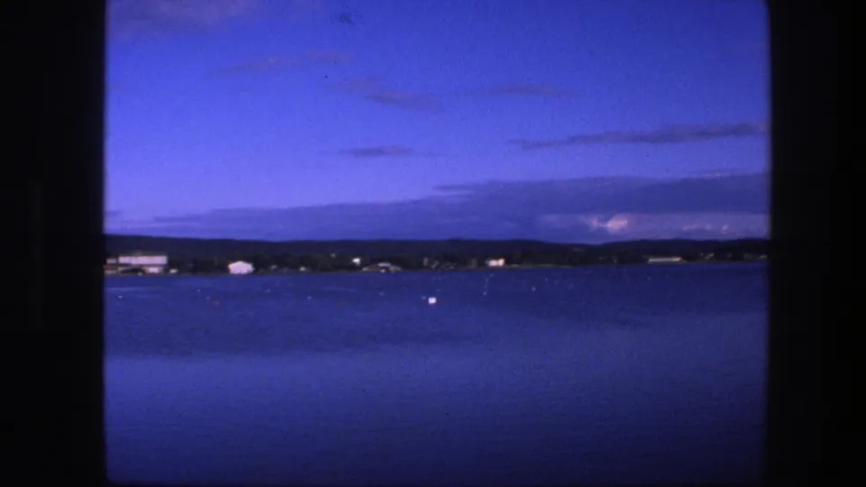 1975 Copenhagen-area 8mm frame from a moving vessel, showing blue water, distant low buildings, a dark shoreline, and cloud bands at dusk.