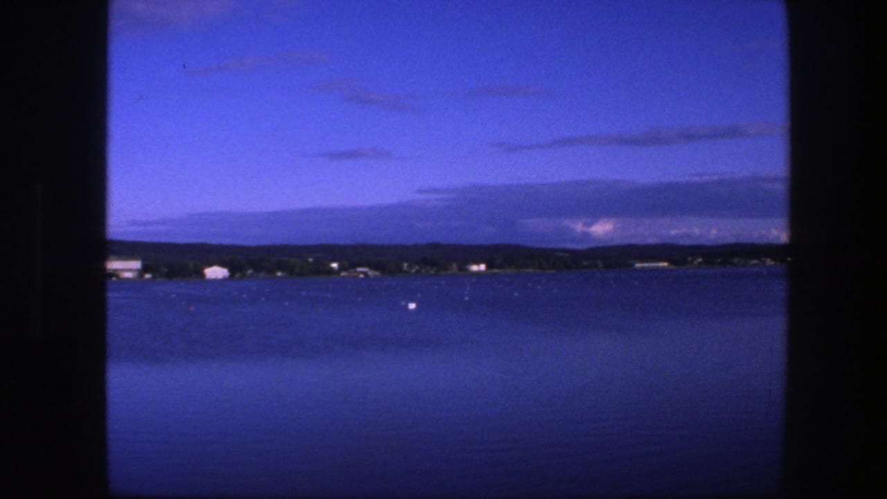1975 Copenhagen home-movie frame showing calm blue water through a dark window frame, with low buildings along the far shore.