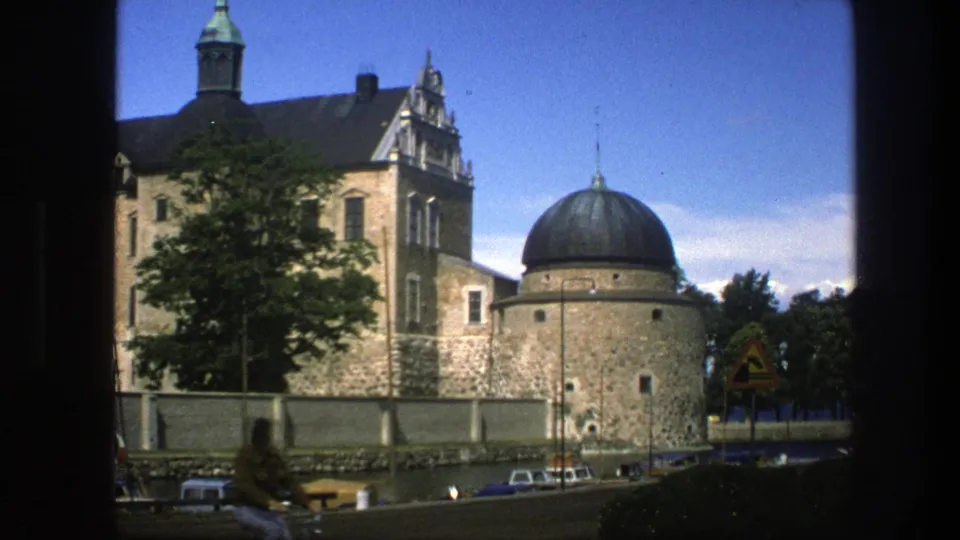 Copenhagen 1975 home-movie view of a stone building, domed round structure, trees, wall, parked cars, and a roadside sign.