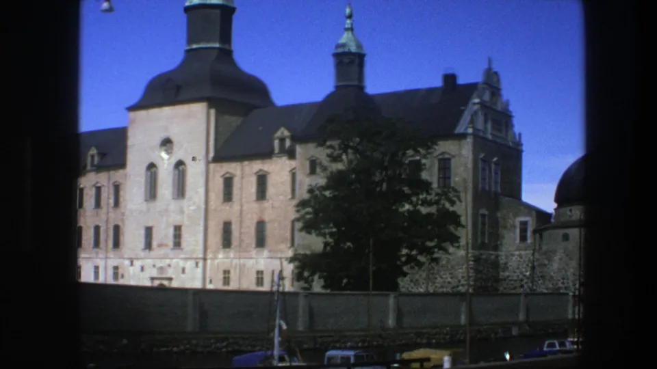 1975 Copenhagen 8mm frame with a closer view of the historic building facade, dark roofline, spire, foreground tree, wall, and cars.