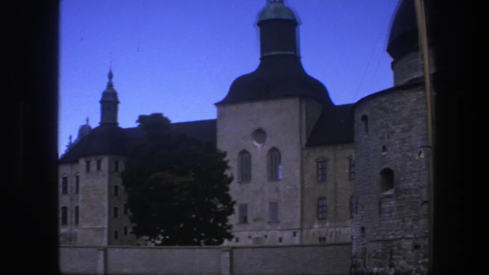 1975 Copenhagen home-movie view of tan historic buildings, tower roofs, arched windows, and a tree framed by a dark opening.