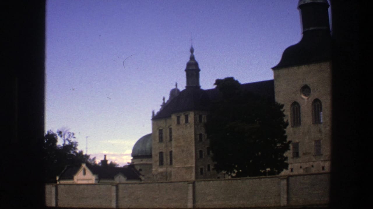 1975 Copenhagen home-movie frame showing a historic towered building, trees, wall, and fading sky through a window frame.