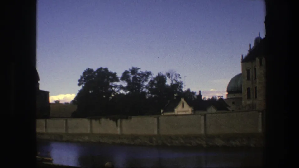 1975 Copenhagen home-movie frame showing a domed stone structure, wall, trees, and water under a blue evening sky.