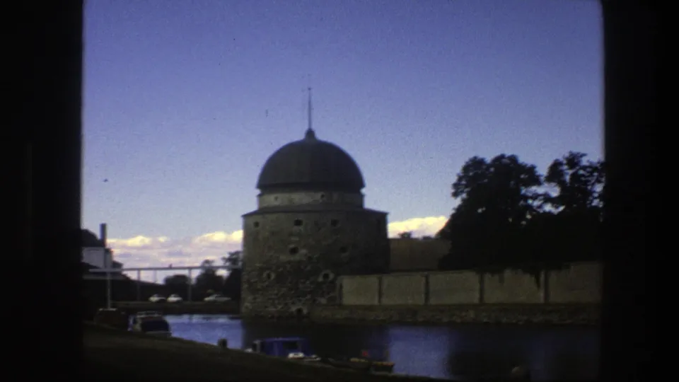 1975 Copenhagen home-movie frame showing a domed waterside structure, parked cars, trees, and dark film-frame edges.