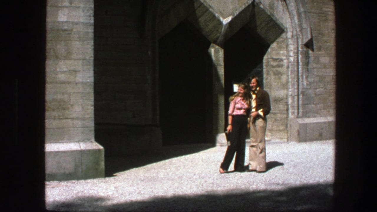 Man and woman stand together in sun outside a stone arched entrance.