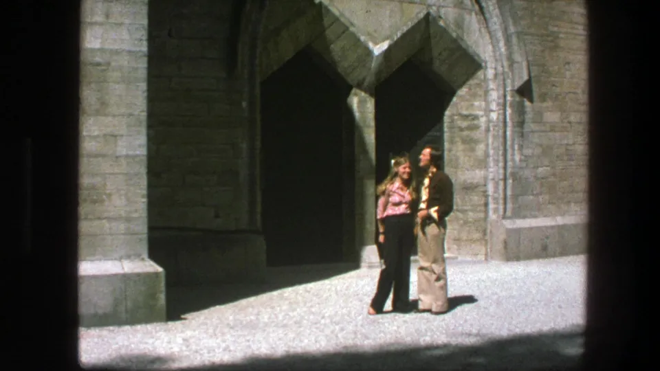 The man and woman remain close together before the historic stone entrance, with bright ground and heavy doorway shadow visible.