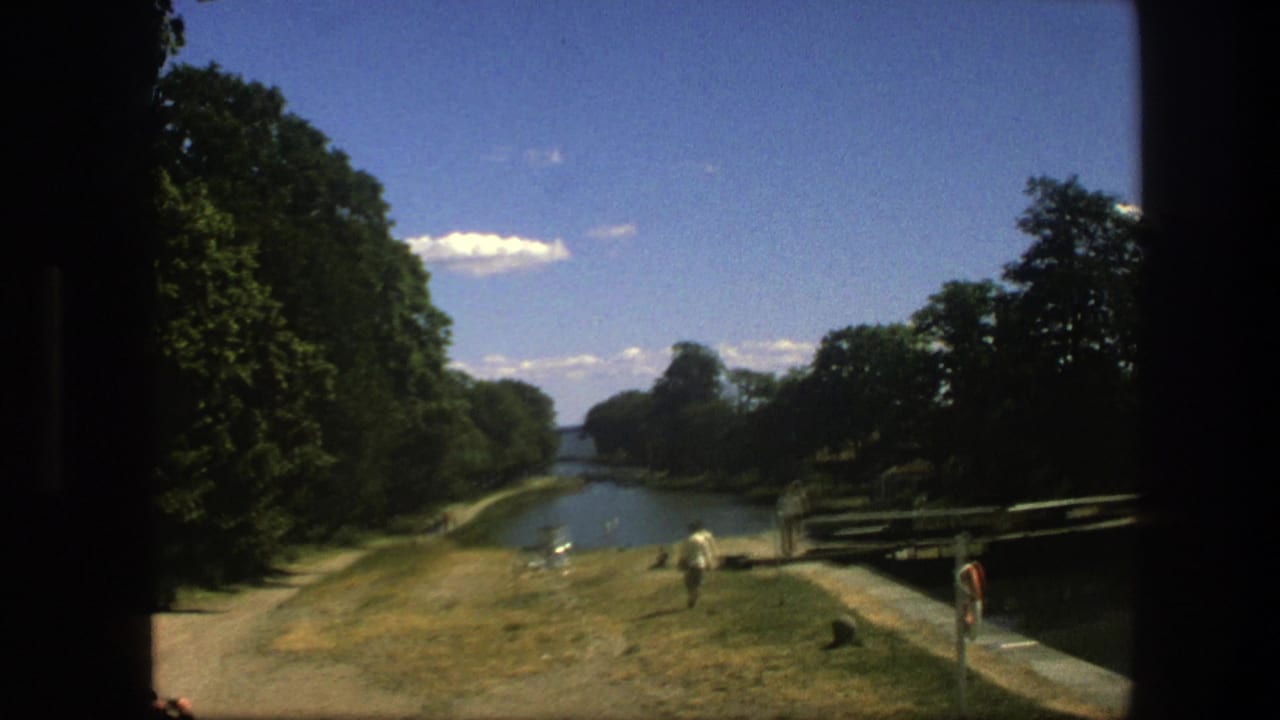 1975 Copenhagen home-movie view of a canal bordered by trees, grass, and a walking path, with a few people visible in the distance.