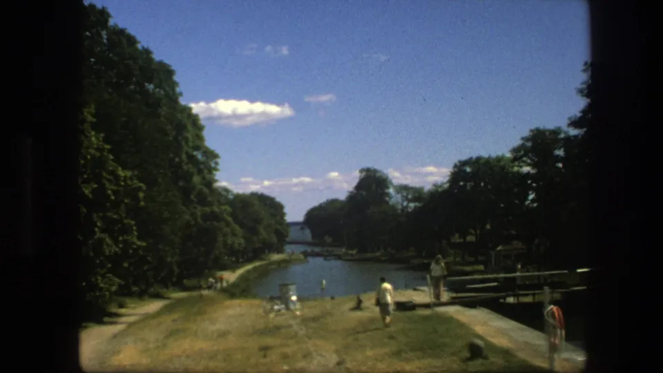 Copenhagen canal-side park scene from 1975, showing people near the water, leafy trees, paths, and a bright sky.