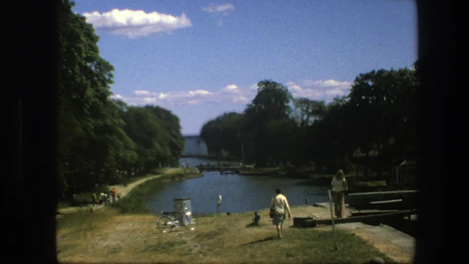 1975 Copenhagen canal view with a bicycle on the grass, pedestrians near the path, dense trees, and water beyond the bank.
