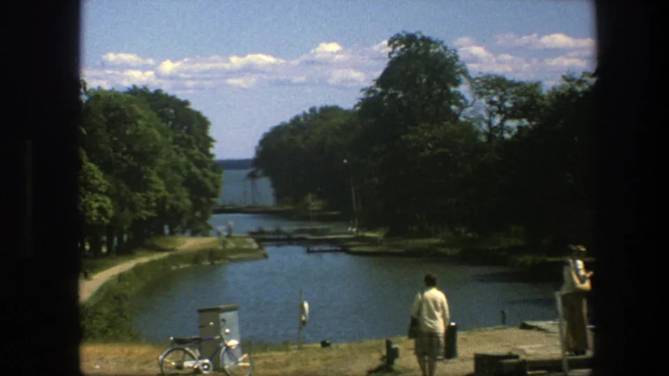 Copenhagen 1975 home-movie frame showing a person walking by the canal, a bicycle nearby, trees, paths, and another person at the side.