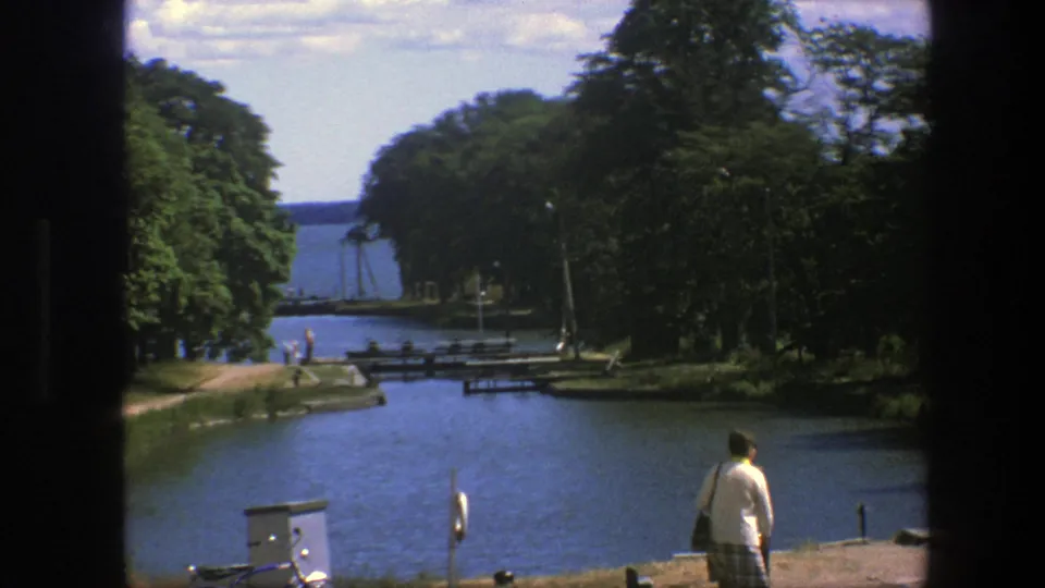 1975 Copenhagen waterfront frame showing an adult near the canal edge, a tree-lined path, distant people, and pier structures by the water.