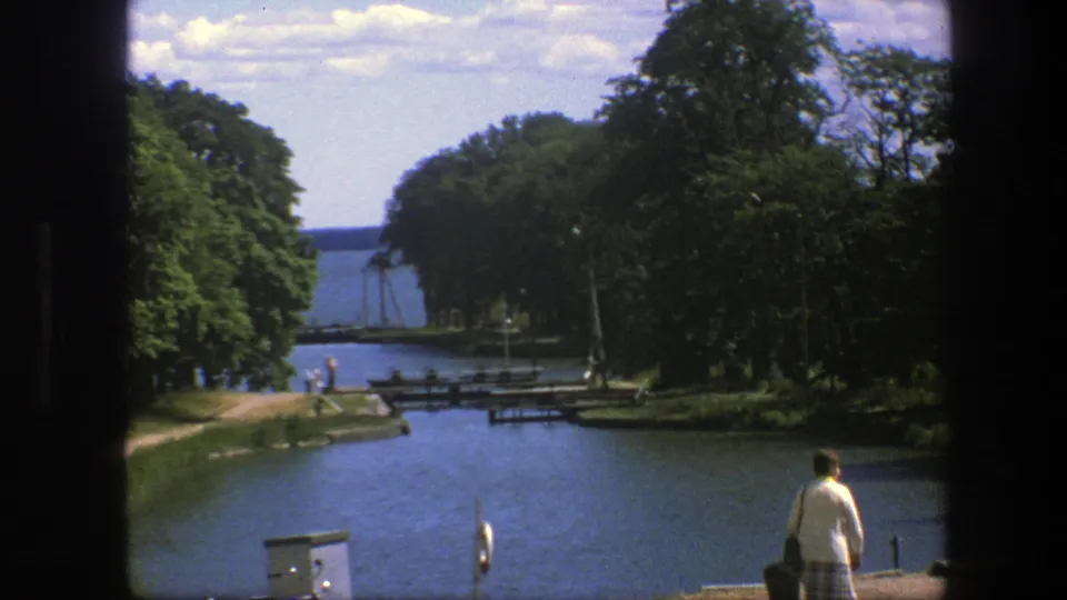 1975 Copenhagen canal scene with a nearby adult figure, distant walkers on the path, wooden pier structures, trees, and open water beyond.
