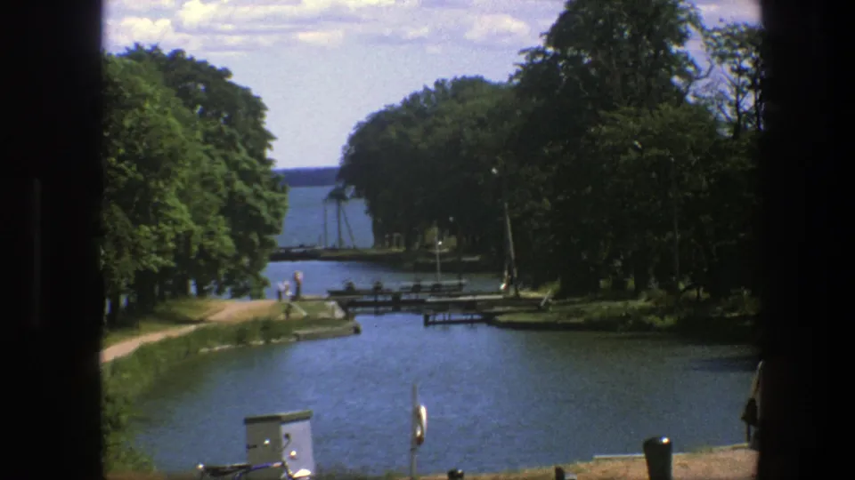 1975 Copenhagen waterfront view with canal water, dense trees, a path, distant pedestrians, pier structures, and a partial adult at the right edge.