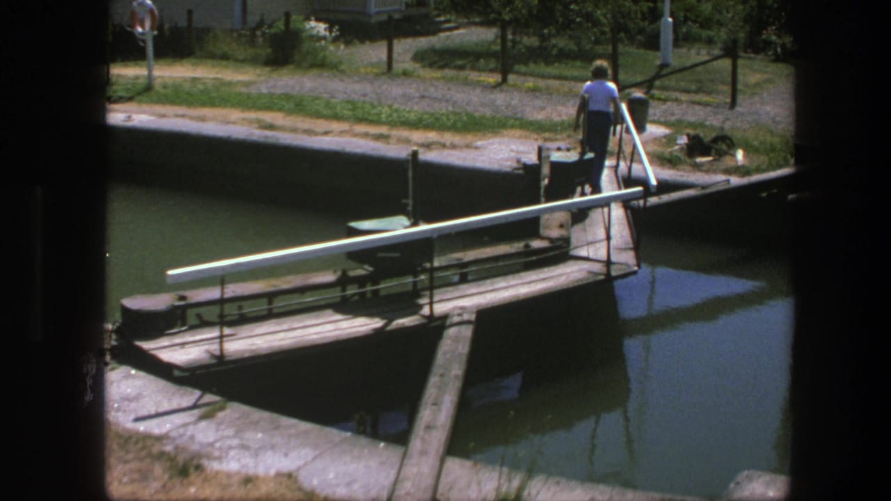 In 1975 Copenhagen footage, a man in a white shirt stands by a narrow bridge over canal water near utility fixtures.