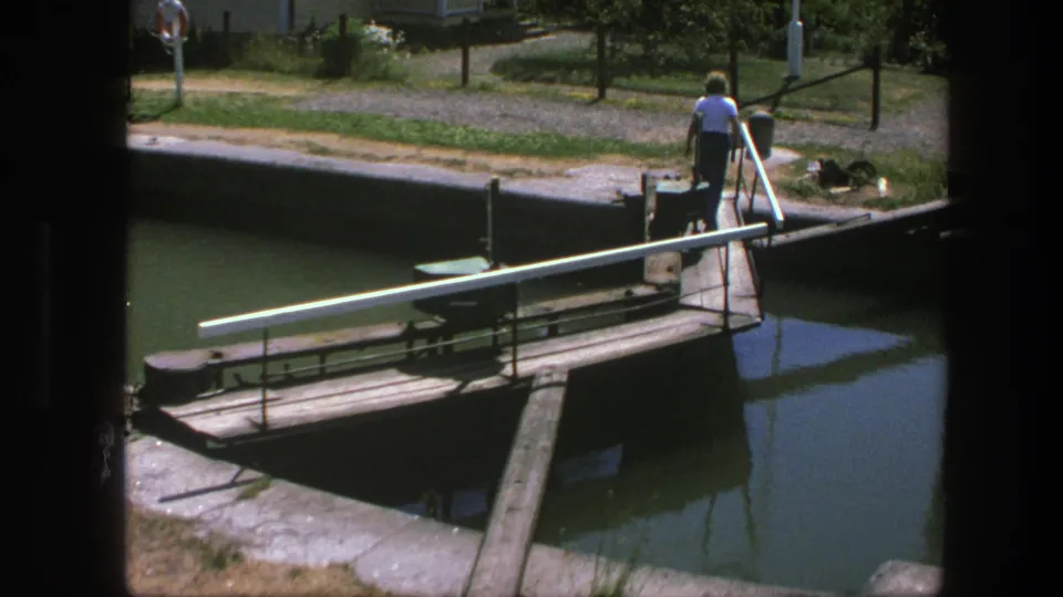 In 1975 Copenhagen footage, a man in a white shirt stands by a narrow bridge over canal water near utility fixtures.